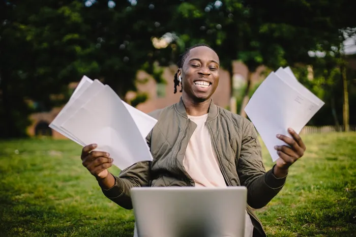 Happy student with documents and a laptop outdoors, celebrating success